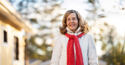 Nainen hymyilee talvisessa maisemassa ulkona. Hänellä on punainen kaulahuivi ja valkoinen takki.