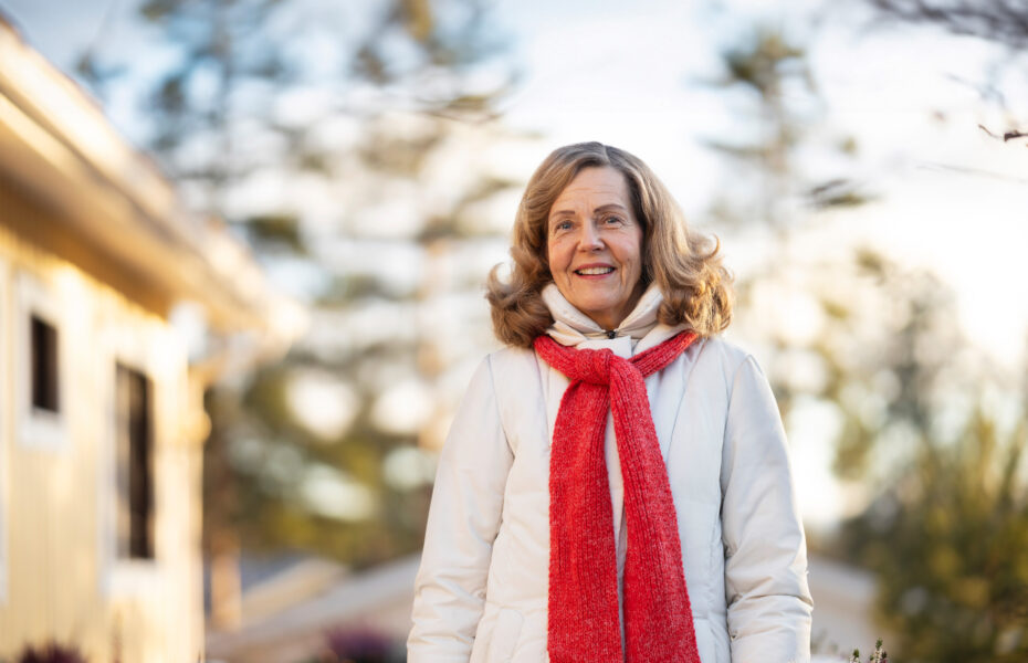 Nainen hymyilee talvisessa maisemassa ulkona. Hänellä on punainen kaulahuivi ja valkoinen takki.