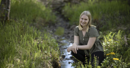 Katri Lemetyinen hymyilee kyykistyneenä pienen puron äärelle saniaisten keskelle.
