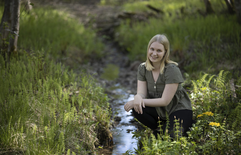 Katri Lemetyinen hymyilee kyykistyneenä pienen puron äärelle saniaisten keskelle.