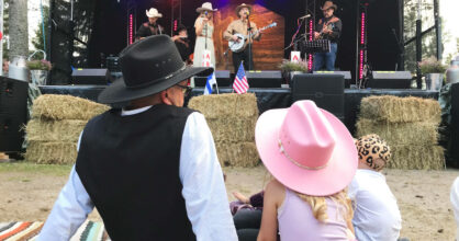 Mies ja pikkutyttö istuvat maassa ja seuraavat lavalla olevaa musiikkiesitystä. Heillä on cowboy-hatut päässä.