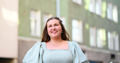 Anni Niskanen seisoo kerrostalon edustalla ja katselee kohti taivasta.