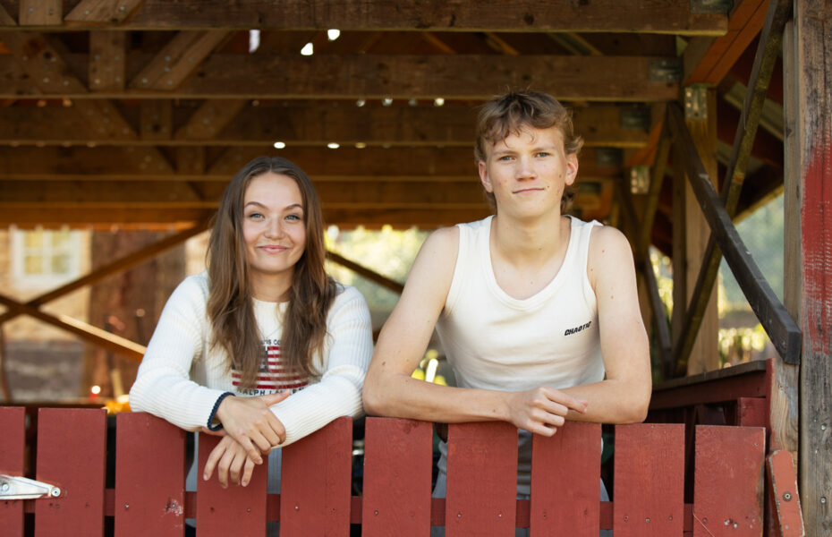 Annika ja Heikki nojailevat punaiseen aitaan. Molemmilla on valkoiset paidat päällään.