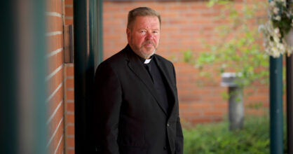 Antti Kruus seisoo mustissa papin vaatteissa punaisen seinän vieressä