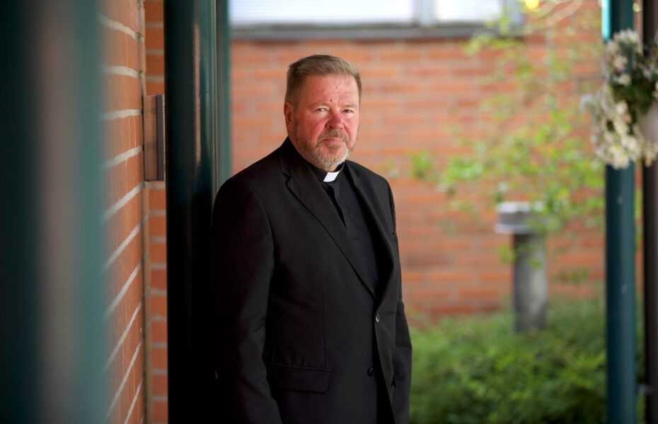 Antti Kruus seisoo mustissa papin vaatteissa punaisen seinän vieressä