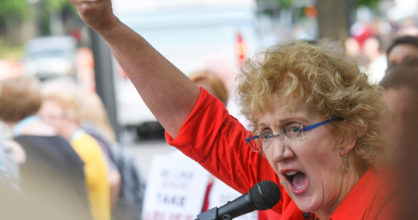 Keski-ikäinen nainen puhuu mikrofoniin ja nostaa toisen kätensä nyrkissä ilmaan. Hänen takanaan on ihmisiä ja katu.
