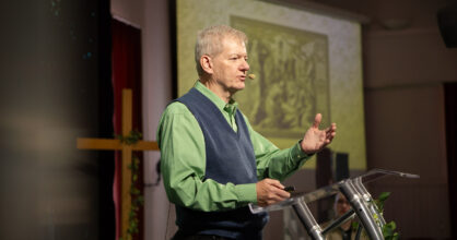 Mies vihreässä kauluspaidassa ja harmaassa liivissä puhujapöntön takana.