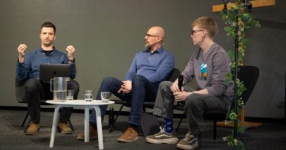 Kolme miestä istuu tuoleilla vierekkäin. Vasemman puoleinen puhuu kädet hieman kohotettuina.