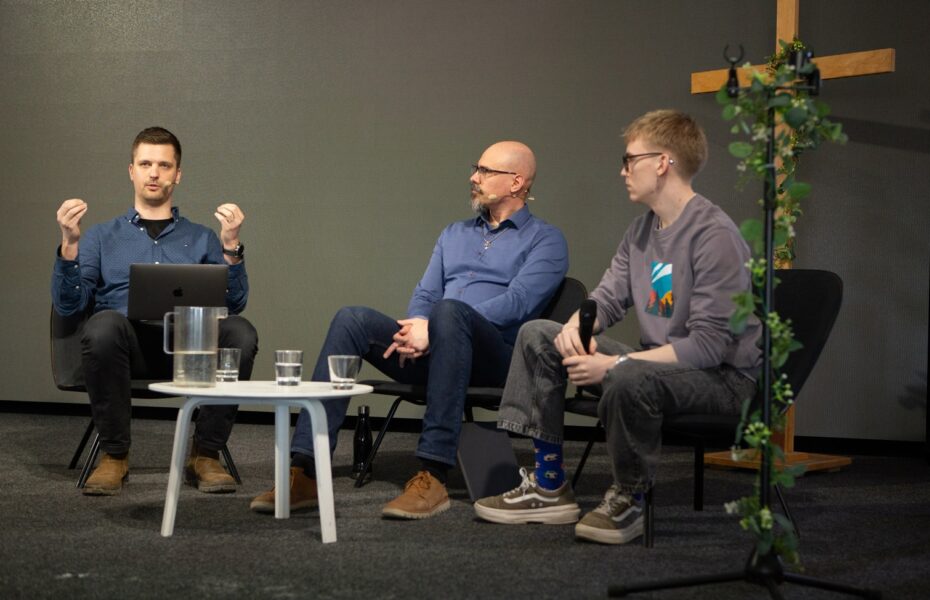 Kolme miestä istuu tuoleilla vierekkäin. Vasemman puoleinen puhuu kädet hieman kohotettuina.