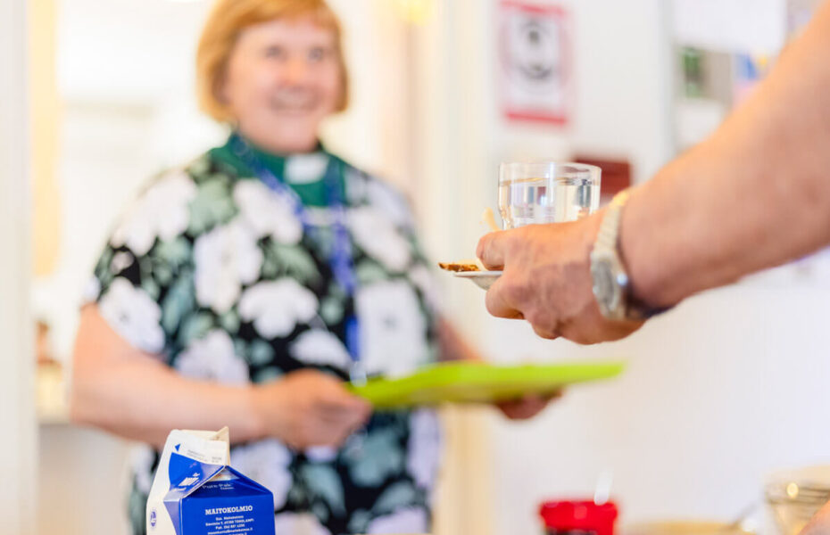 Diakoniatyöntekijä tarjoaa ruokaa miehelle, joka pitää kädessään lasia.