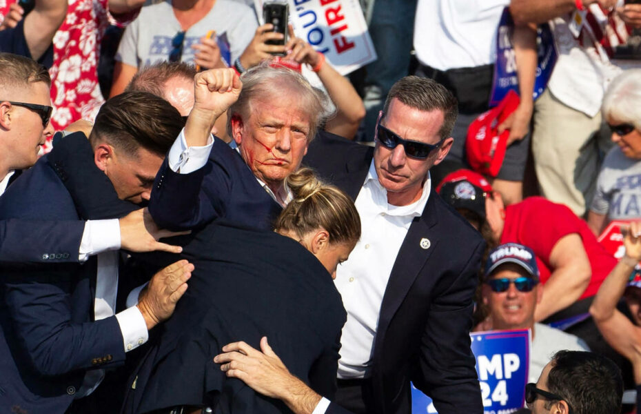 Donald Trump on nostanut nyrkkinsä ilmaan. Hänen poskellaan on verta. Tummiin pukeutuneet ihmiset suojaavat häntä.