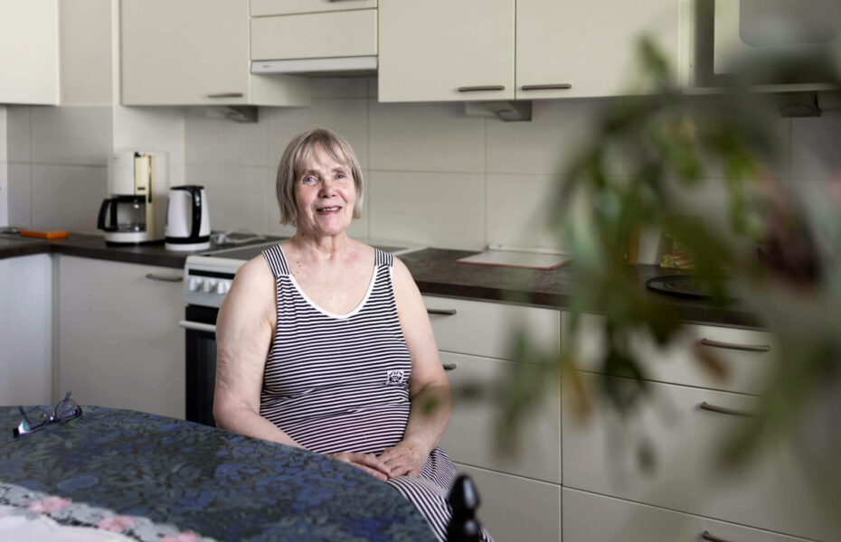 Kuvassa Eeva Piiroinen istuu keittiön pöytänsä ääressä ja hymyilee.