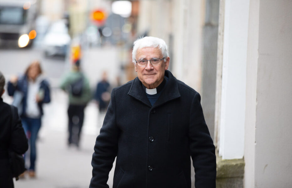 Harmaantunut ja silmälasipäinen mies, jolla on musta takki ja papin panta.