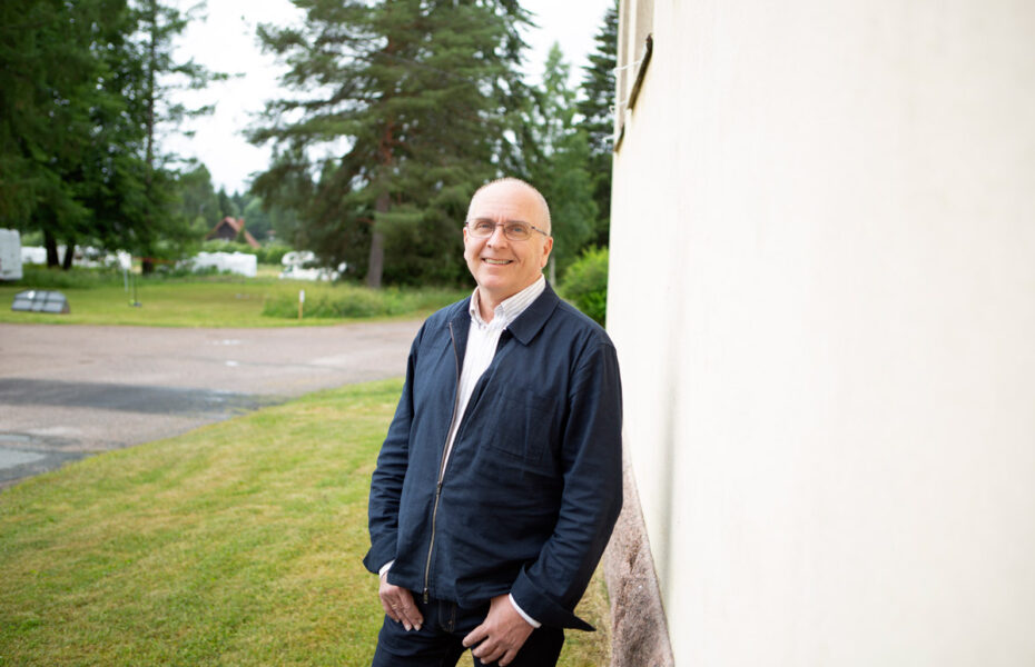 Kaljupäinen ja silmälasipäinen Gunnar seisoo vaalean seinän vieressä nurmikolla.