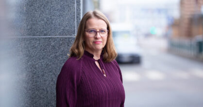 Hanna Salomäki hymyilee. Hänellä on silmälasit ja violetti paita sekä hartioille asti ulottuvat hiukset.