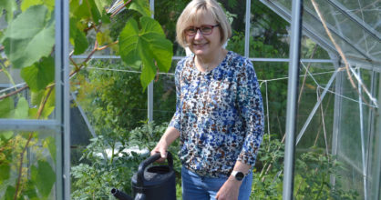 Hannele Karppinen seisoo pienessä kasvihuoneessa kastelukannu kädessään.