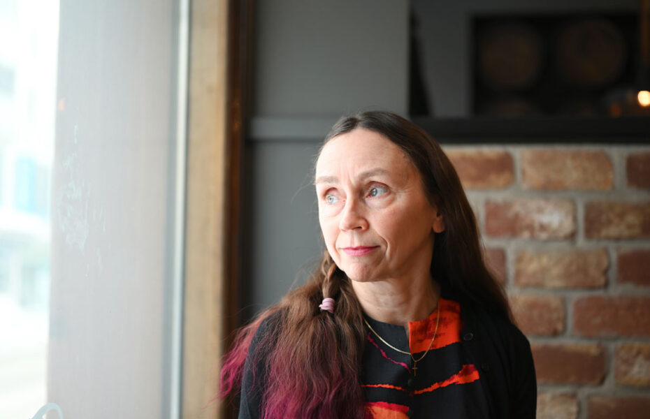 Keski-ikäinen nainen katsoo ikkunasta ulos. Hänellä on pitkät ruskeat hiukset, joiden latvat on värjätty violetiksi.