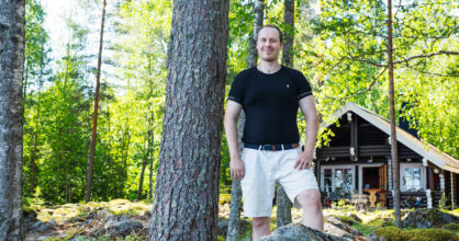 Heikki poseeraa metsässä hirsimökki taustalla