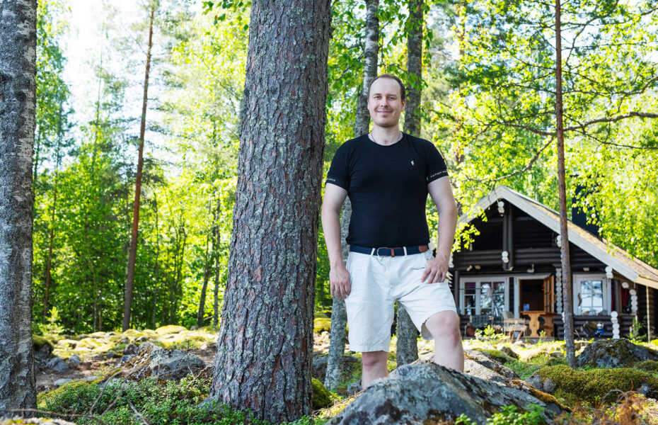 Heikki poseeraa metsässä hirsimökki taustalla