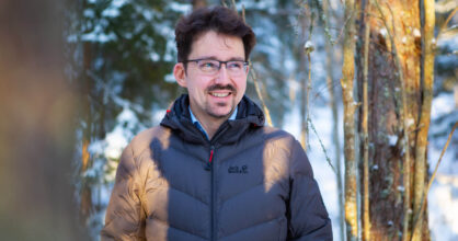 Silmälasipäinen ja mustahiuksinen mies hymyilee lumisessa metsässä.