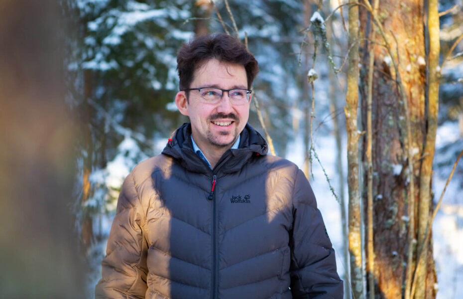 Silmälasipäinen ja mustahiuksinen mies hymyilee lumisessa metsässä.