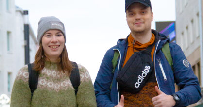 Nainen ja mies hymyilee kaupungin kadulla. Miehellä on peukalot pystyssä.