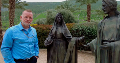 Siniseen kauluspaitaan pukeutunut mies seisoo kahden patsaan vieressä.