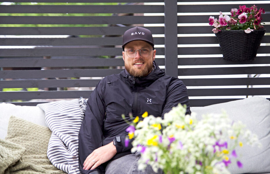 Joakim istuu terassilla sohvalla. Hänellä on lippalakki ja silmälasit.