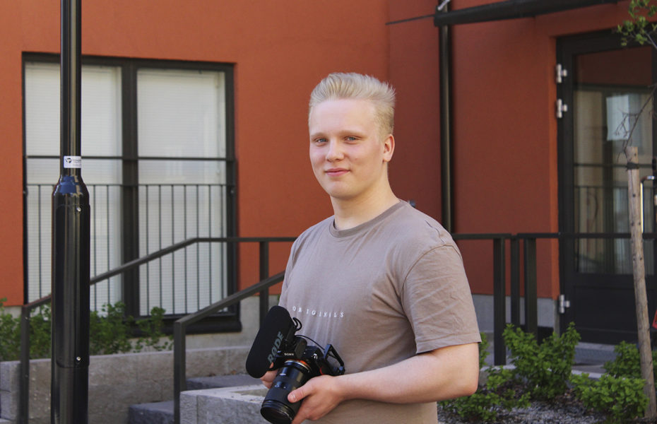 Joel Tiainen seisoo kerrostalon edustalla. Hän pitää käsissään kameraa.