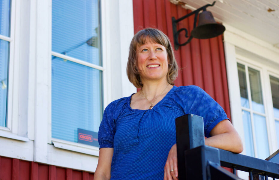 Johanna Sandberg nojaa portaan kaiteeseen ja katsoo kaukaisuuteen. Hänellä on polkkahiukset ja sininen paita.