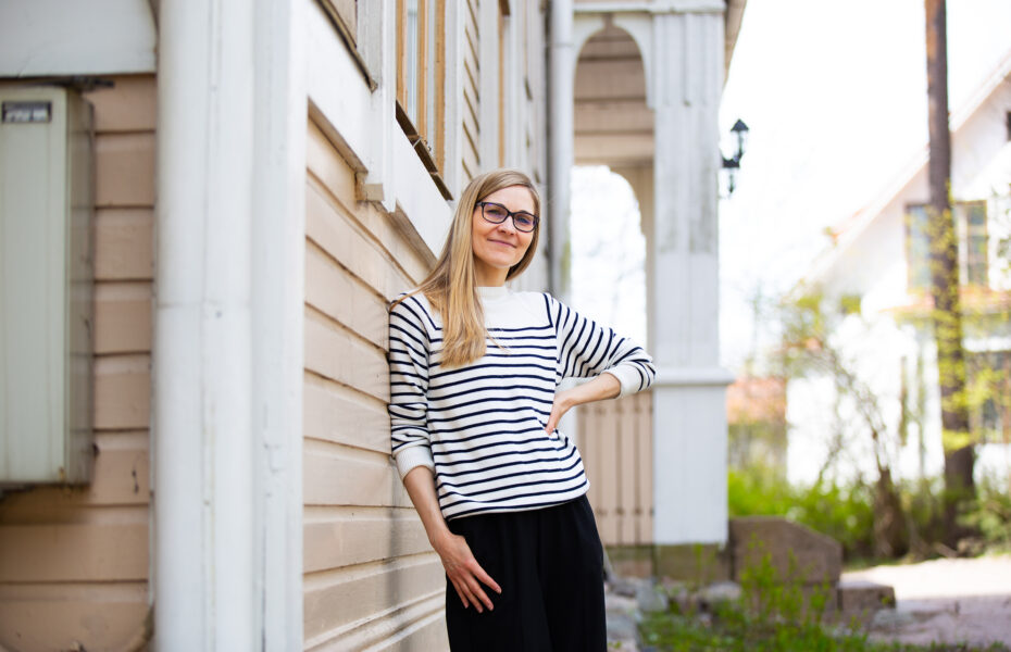 Nainen nojaa talon seinään ja hymyilee. Naisella on pitkät hiukset ja mustat silmälasit.