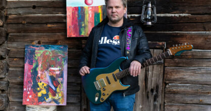 JP Löfberg seisoo kitaran kanssa hirsiseinän edessä.