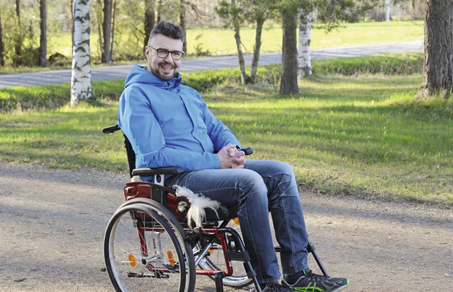 Juha Vihriälä istuu pyörätuolissa pihatiellä. Hänen taakseen jää puita ja nurmikkoa.