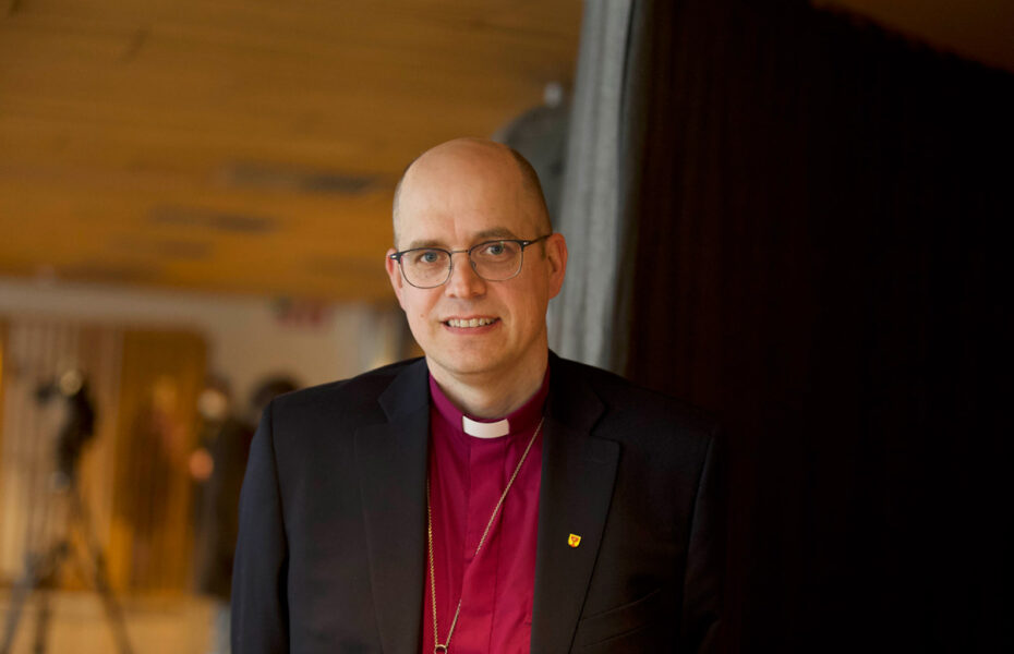 Kalju ja silmälasipäinen mies hymyilee. Hänellä on musta puvun takki ja sen alla violetti papinpaita.