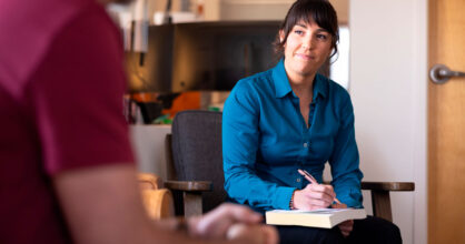 Nainen istuu nojatuolissa ja kirjaa ylös paperiin samalla, kun hän kuuntelee miestä.