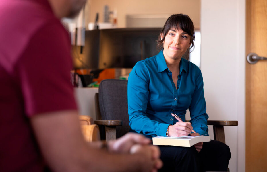Nainen istuu nojatuolissa ja kirjaa ylös paperiin samalla, kun hän kuuntelee miestä.