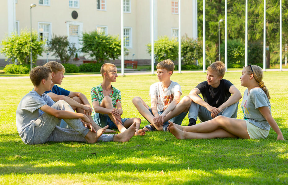Viisi poikaa ja yksi tyttö istuvat puolikaaressa nurmikolla kesäisenä päivänä. Heidän takanaan on Kansanlähetysopisto.