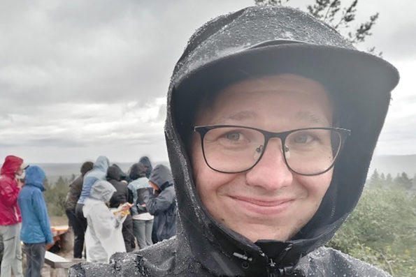 Antti Koskenniemi hymyilee kastunut sadetakki yllään mäen päällä. Hänen takanaan on sadevaatteisiin pukeutuneita hahmoja sekä kaukana näkyvä horisontti.