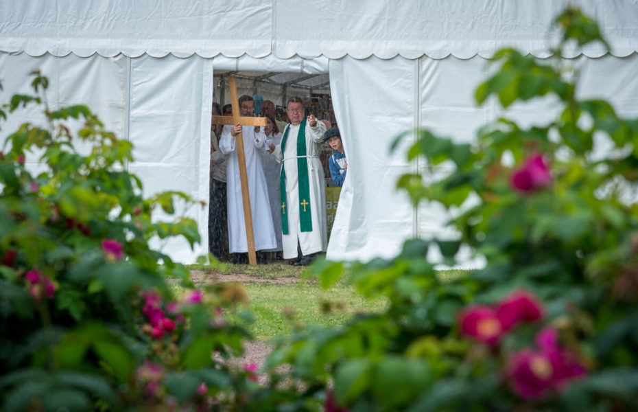 Pappi ja muu jumalanpalveluksen ristikulkue katsovat ulos juhlateltasta sateeseen. Myös nuori poika kurkistaa teltan seinän takaa.