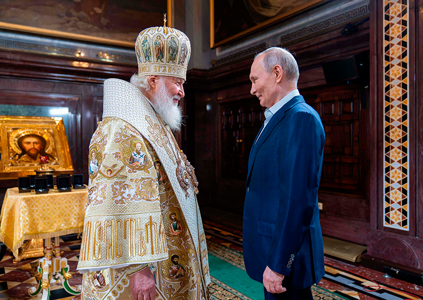 Kaksi miestä seisoo vastakkain. Vasemmalla puolella olevalla on patriarkan kuninkaalliselta näyttävä asu. Oikealla puolelle on Putin sinisessä puvussaan.