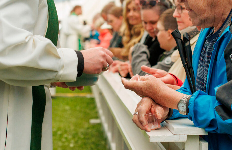 Pappi antaa ehtoollista miehelle, jonka käsi on ojennettu. Ehtoollisenviettäjät ovat suuressa teltassa.