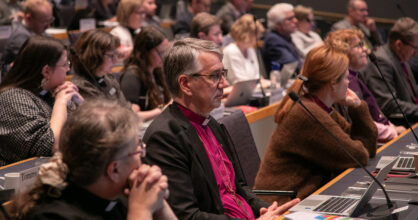 Kuvassa on joukko ihmisiä osallistumassa konferenssiin. Etualalla näkyy mies, joka näyttää miettivältä, ja taustalla on useita muita osallistujia.