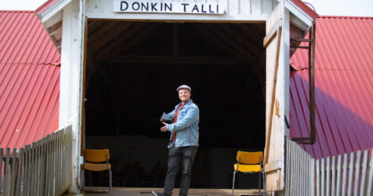 Mies osoittaa kutsuvasti käsiään kohti tallia, jonka yläpuolella on "Donkin talli".