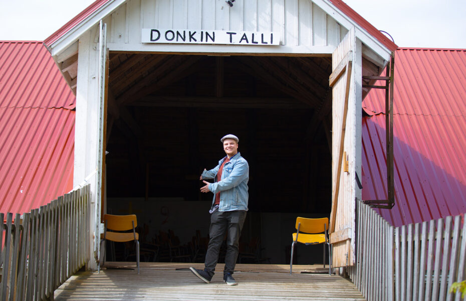 Mies osoittaa kutsuvasti käsiään kohti tallia, jonka yläpuolella on "Donkin talli".