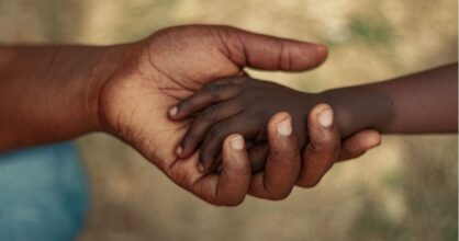 Lapsen käsi aikuisen kädessä. Heillä molemmilla on tumma iho.