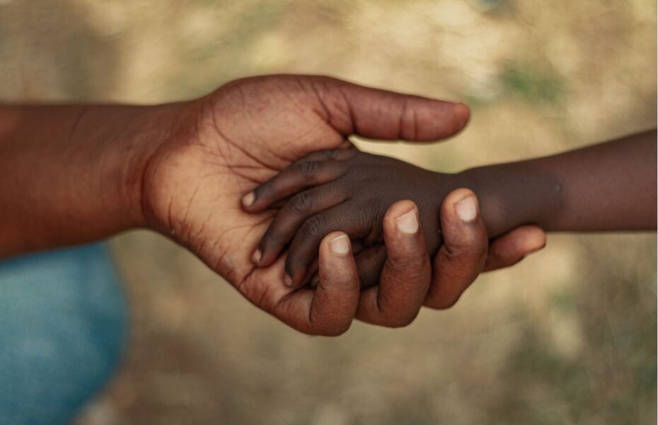 Lapsen käsi aikuisen kädessä. Heillä molemmilla on tumma iho.
