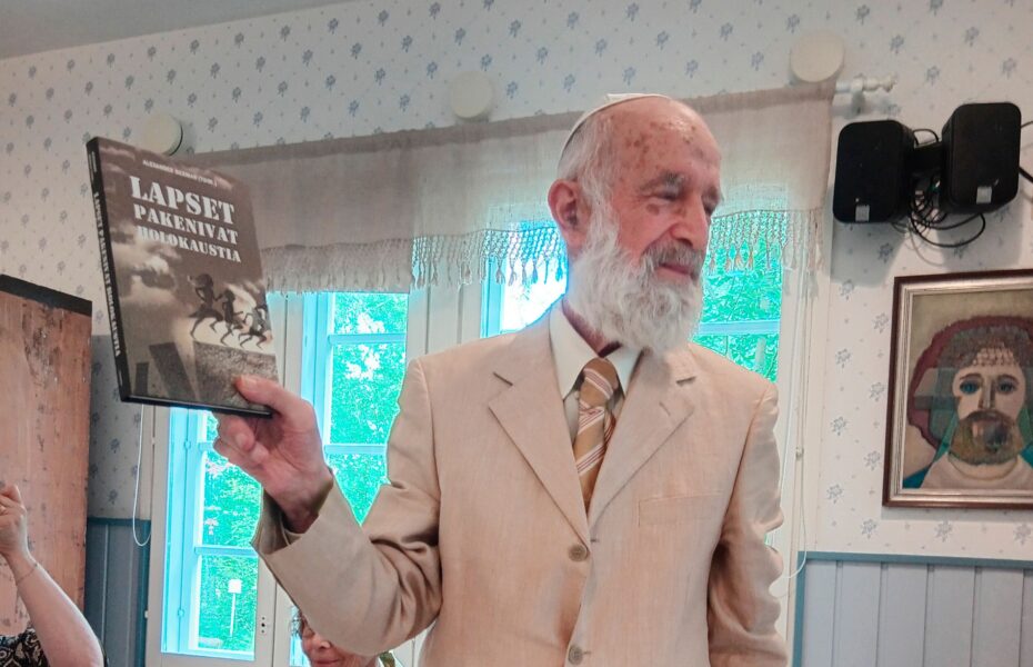 Valkopartainen mies pitää kädessään kirjaa, jossa lukee: "Lapset pakenivat holokaustia".