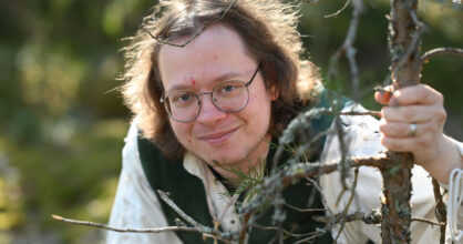 Miehellä on silmälasit ja olkapäille ulottuvat kiharat hiukset. Hän hymyilee.