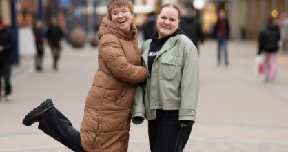 Kaksi nuorta naista hymyilee katukuvassa takit päällä. Vasemmanpuolisella on jalka nostettu koukkuun ilmaan.