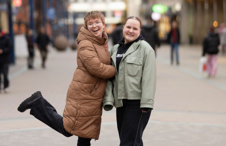 Kaksi nuorta naista hymyilee katukuvassa takit päällä. Vasemmanpuolisella on jalka nostettu koukkuun ilmaan.